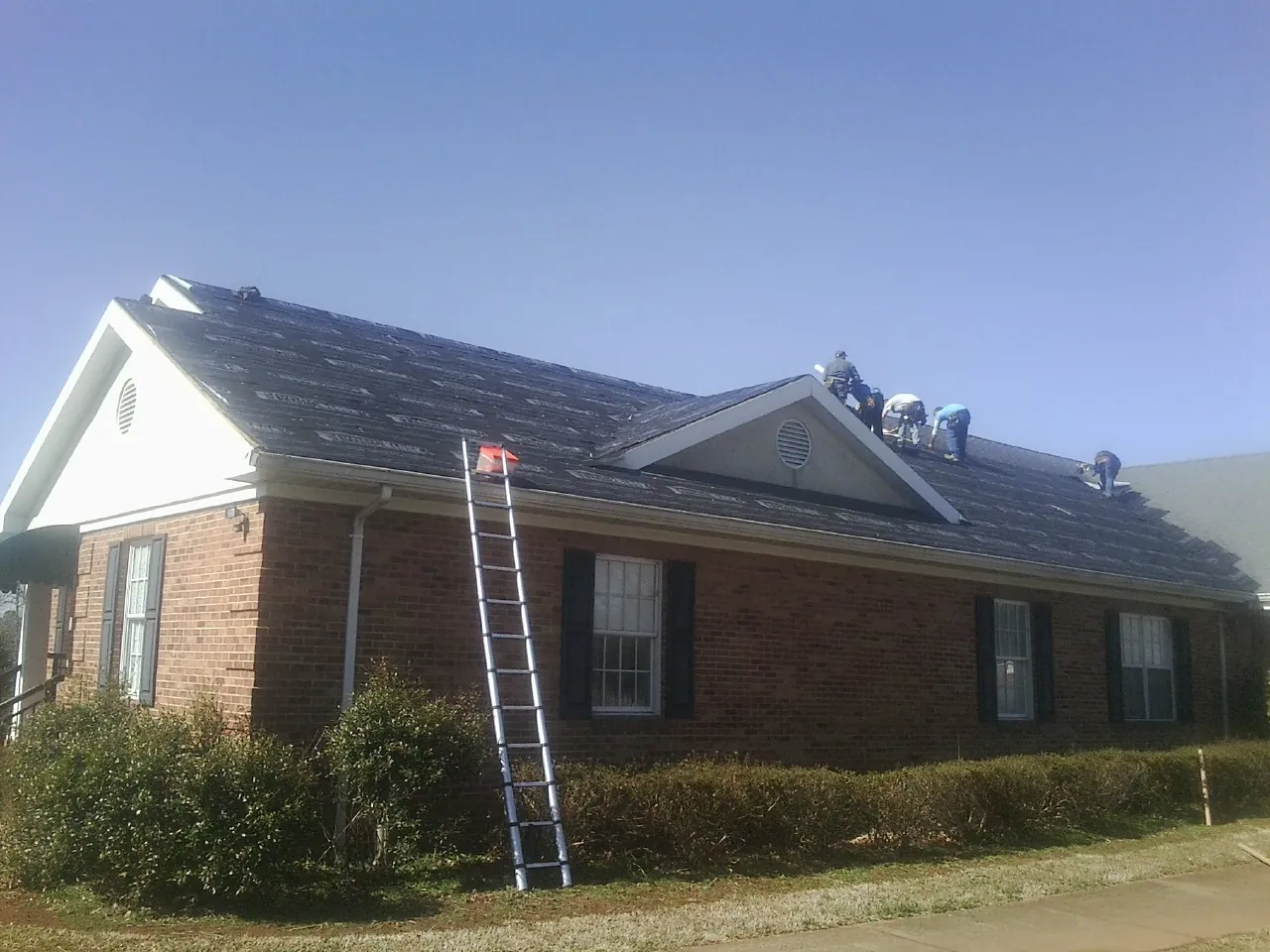 Crew tearing shingles off a brick ranch roof with a ladder set — Emergency Roof Repair work by our crew in Kirkman