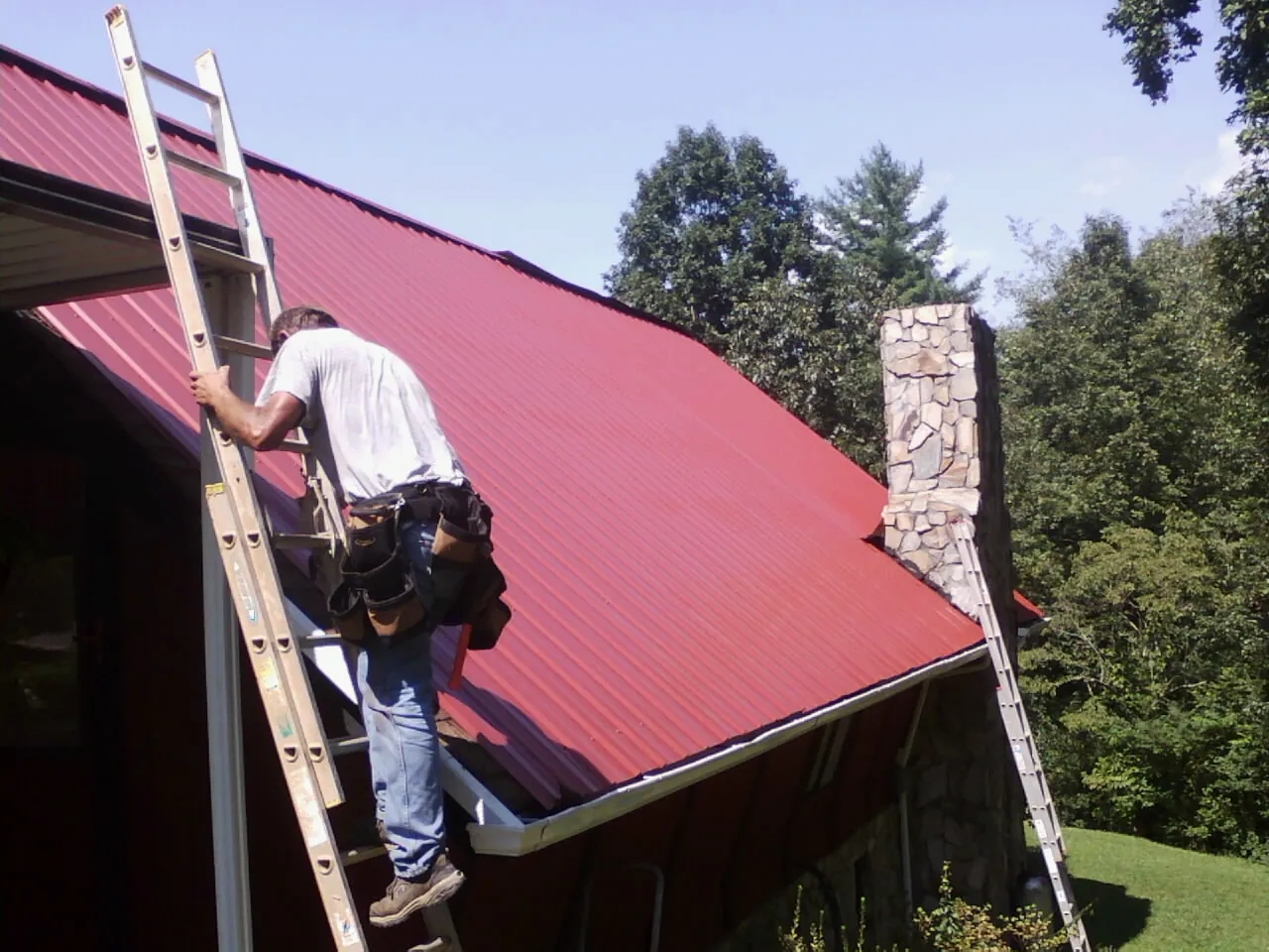 Roofer on a ladder beside a red metal roof and stone chimney — Soffit & Fascia Repair by Roofing Brothers in Kirkman